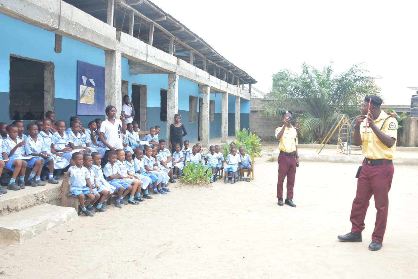 Pupils seated outdoors in the school yard listen to two LASTMA officers