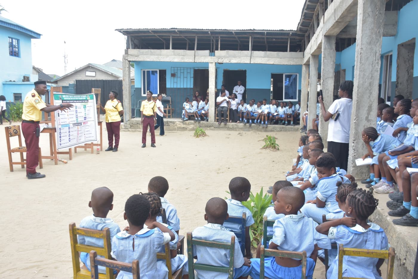 Young pupils seated in the school yard watch LASTMA officers present road safety signs