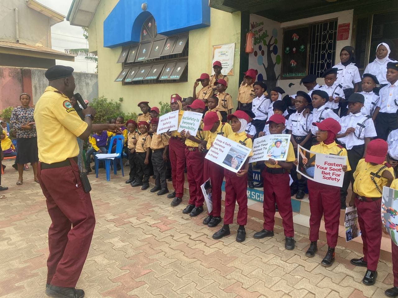 Pupils in Traffic Safety Advocacy Club uniform hold road safety placards while a LASTMA officer speaks