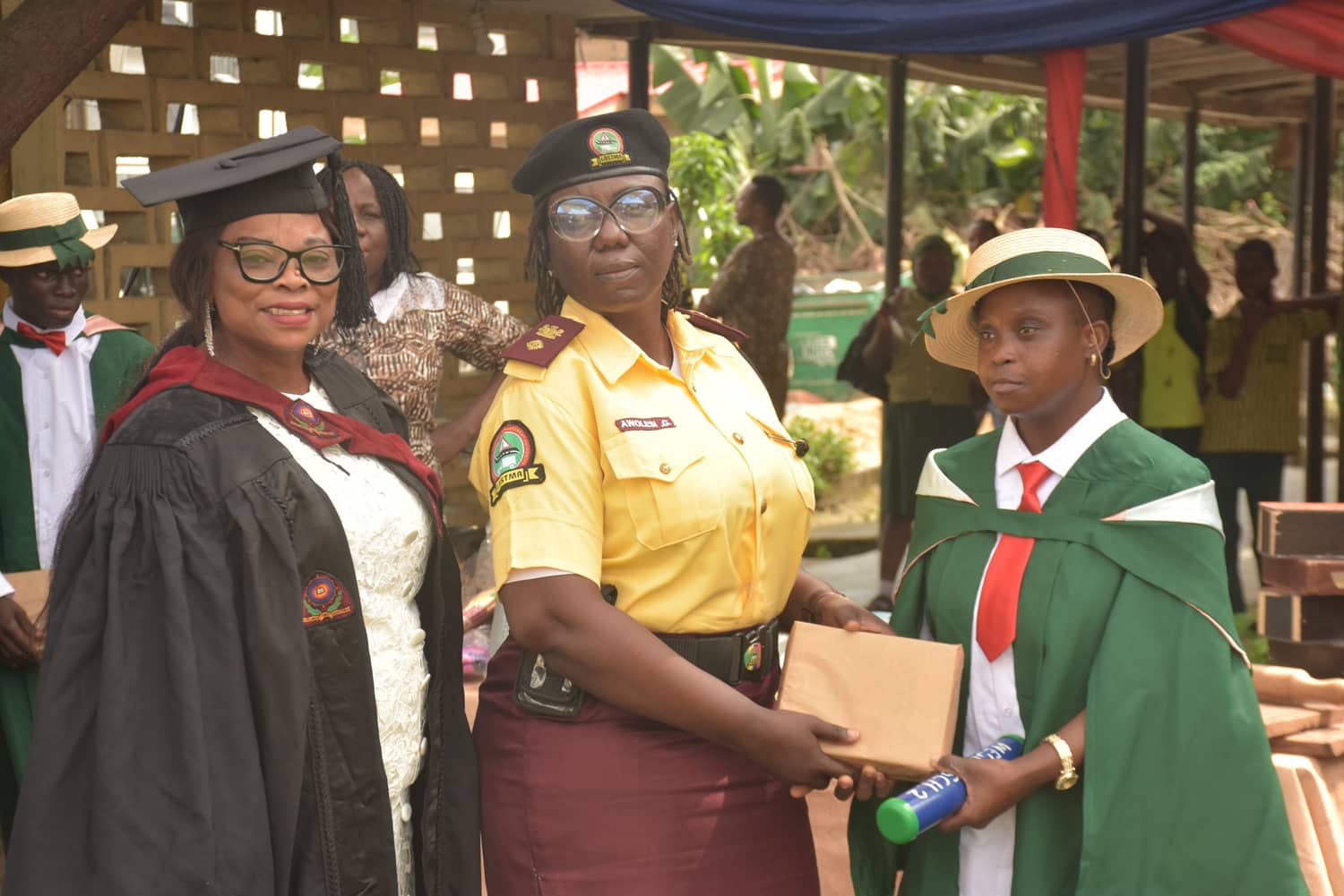 A LASTMA officer presents a prize to a graduating pupil while the school's head, in academic gown, stands with them