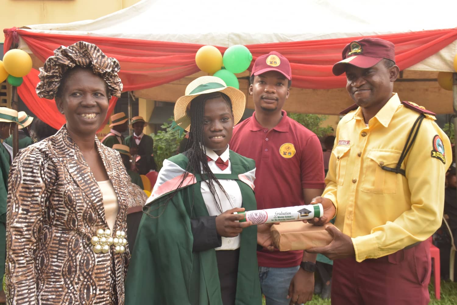 A LASTMA officer hands a graduating girl her certificate and a wrapped prize while her head teacher looks on