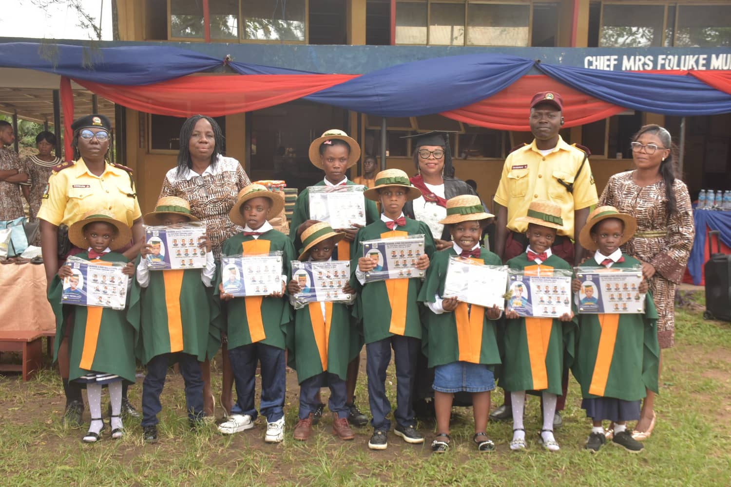 Young pupils in green and orange gowns and straw boaters hold up framed photographs of their class, flanked by two LASTMA officers