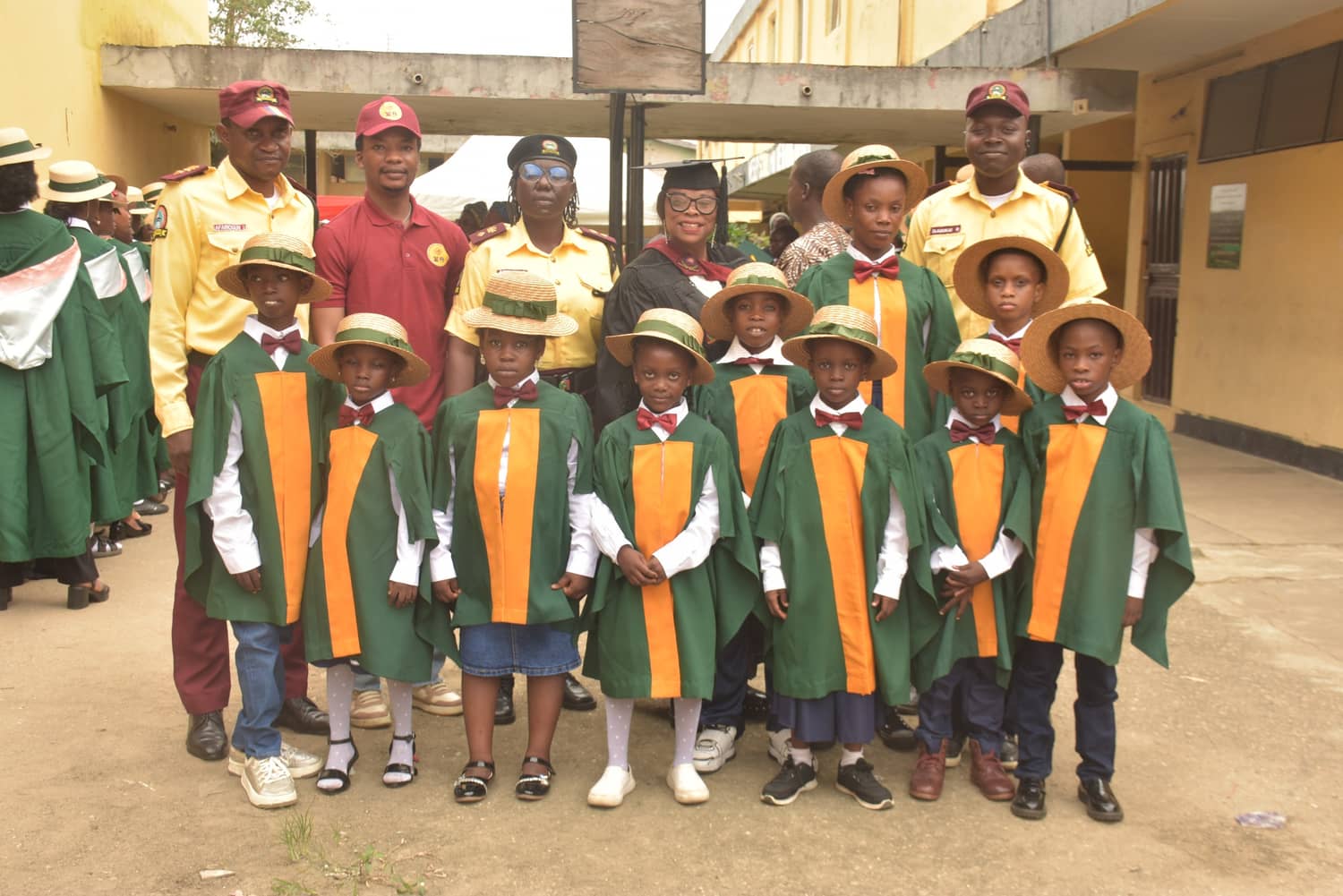 Primary graduates in green and orange gowns stand with three LASTMA officers and their head teacher in the school yard
